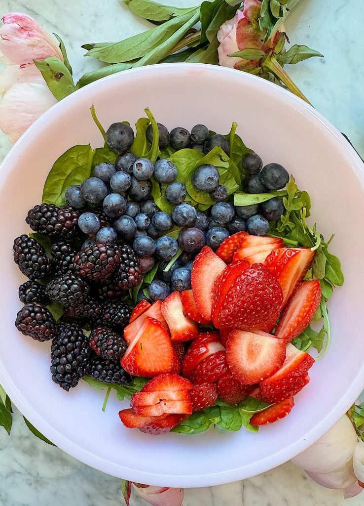 A white bowl containing fresh spinach, blueberries, blackberries, and sliced strawberries on a white surface with peony flowers in the background.