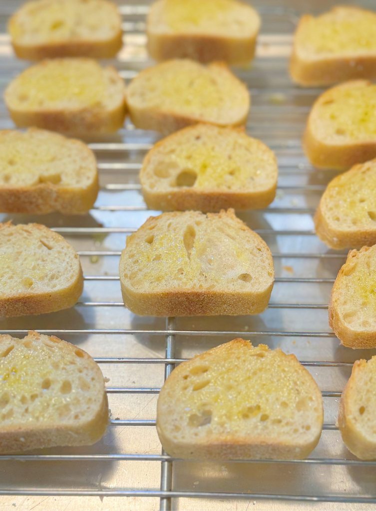Multiple slices of toasted bread with a light drizzle of olive oil are arranged on a cooling rack.