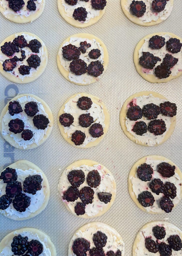 Unbaked cookies topped with dark berries and a creamy mixture, arranged in rows on a baking sheet.