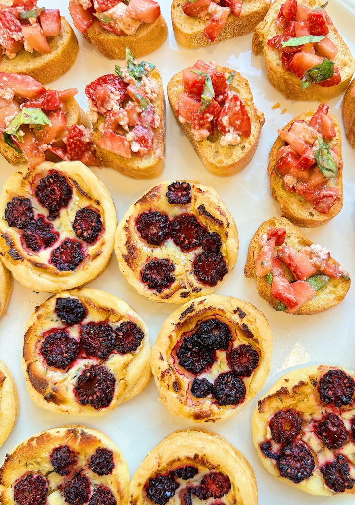 An assortment of bruschetta topped with diced strawberries and basil, alongside mini pastries with a blackberry filling on a white surface.