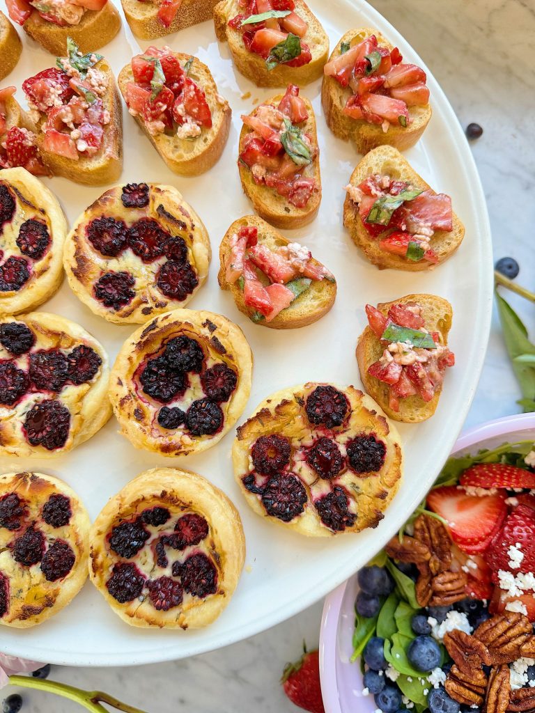 A white platter with blackberry tartlets and bruschetta topped with diced strawberries and herbs. A bowl of salad with berries, pecans, and feta is visible on the side.