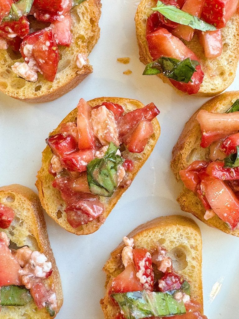 Close-up of bruschetta topped with diced strawberries, fresh basil leaves, and crumbled feta cheese on slices of toasted baguette.