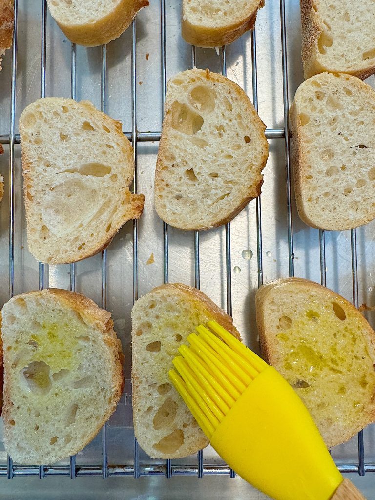 Slices of bread on a cooling rack being brushed with olive oil using a yellow brush.