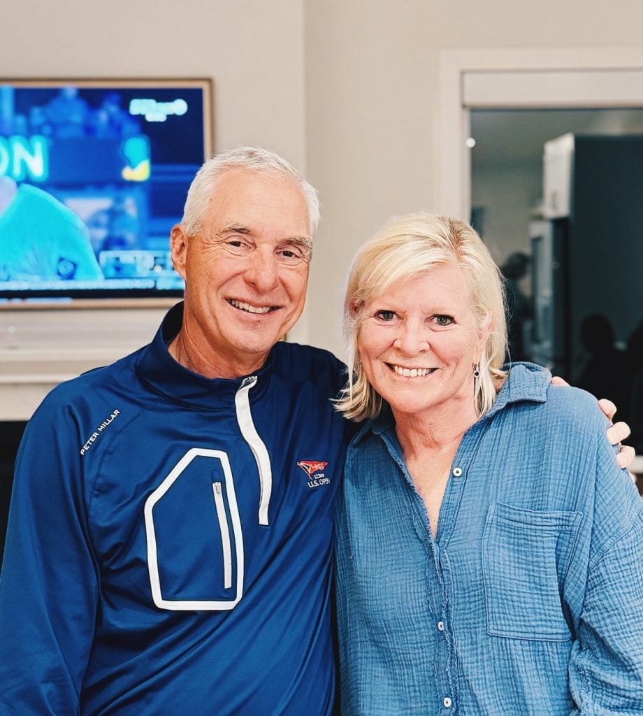 A smiling older man and woman stand together indoors, with a baseball game on TV in the background.