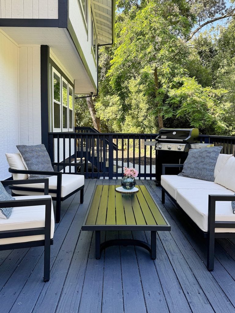 A wooden outdoor deck features a seating area with two cushioned chairs and a sofa, a rectangular table with flowers, and a gas grill in the background. Trees provide a natural backdrop.