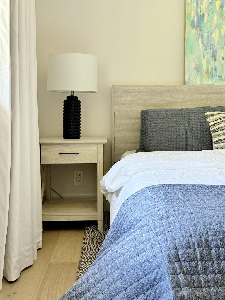 A bedroom with a wooden nightstand, black lamp, and grey and striped pillows on a bed with a grey and blue blanket. A painting with blue and green tones hangs on the wall. White curtains are to the left.