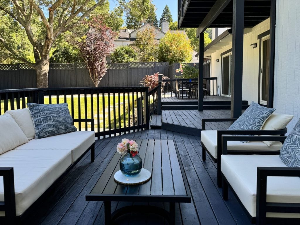 Outdoor patio with black railings, dark wood decking, white cushioned seating, a black coffee table with a vase and flowers, and a view of a green yard and trees in the background.
