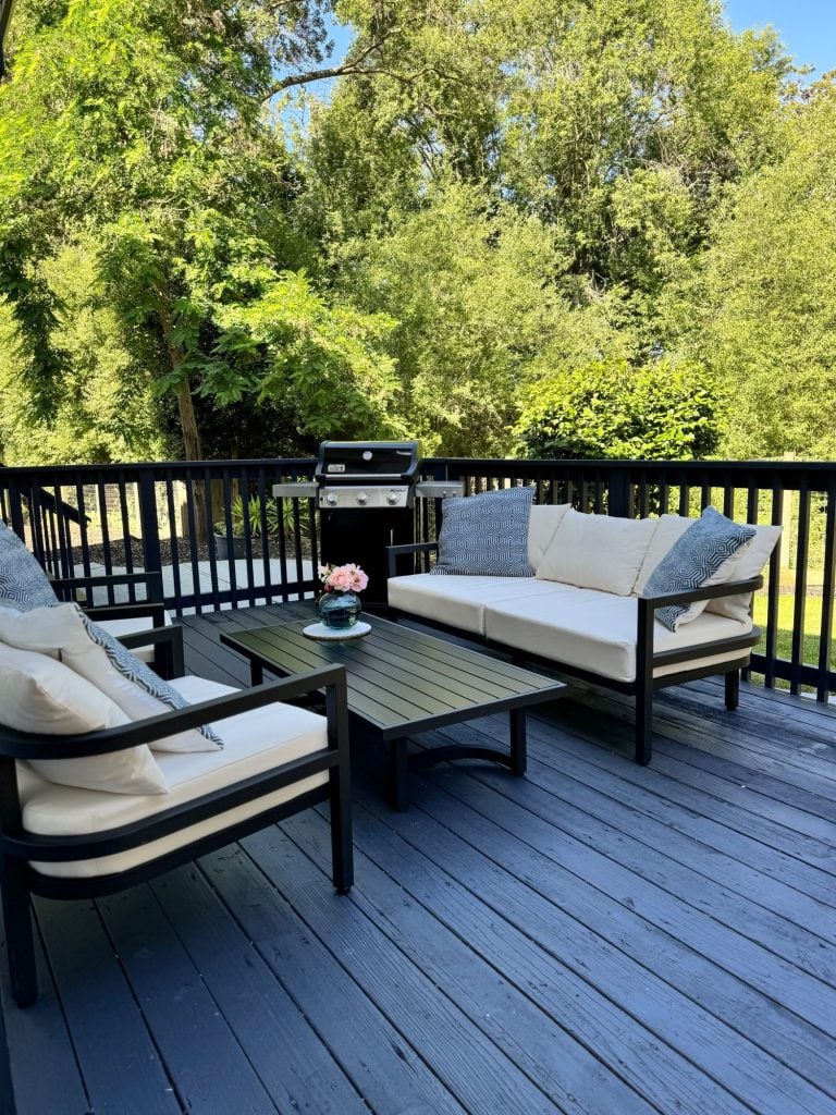 A wooden deck with two cushioned sofas, a low table, a small flower arrangement, and a gas grill, surrounded by greenery.