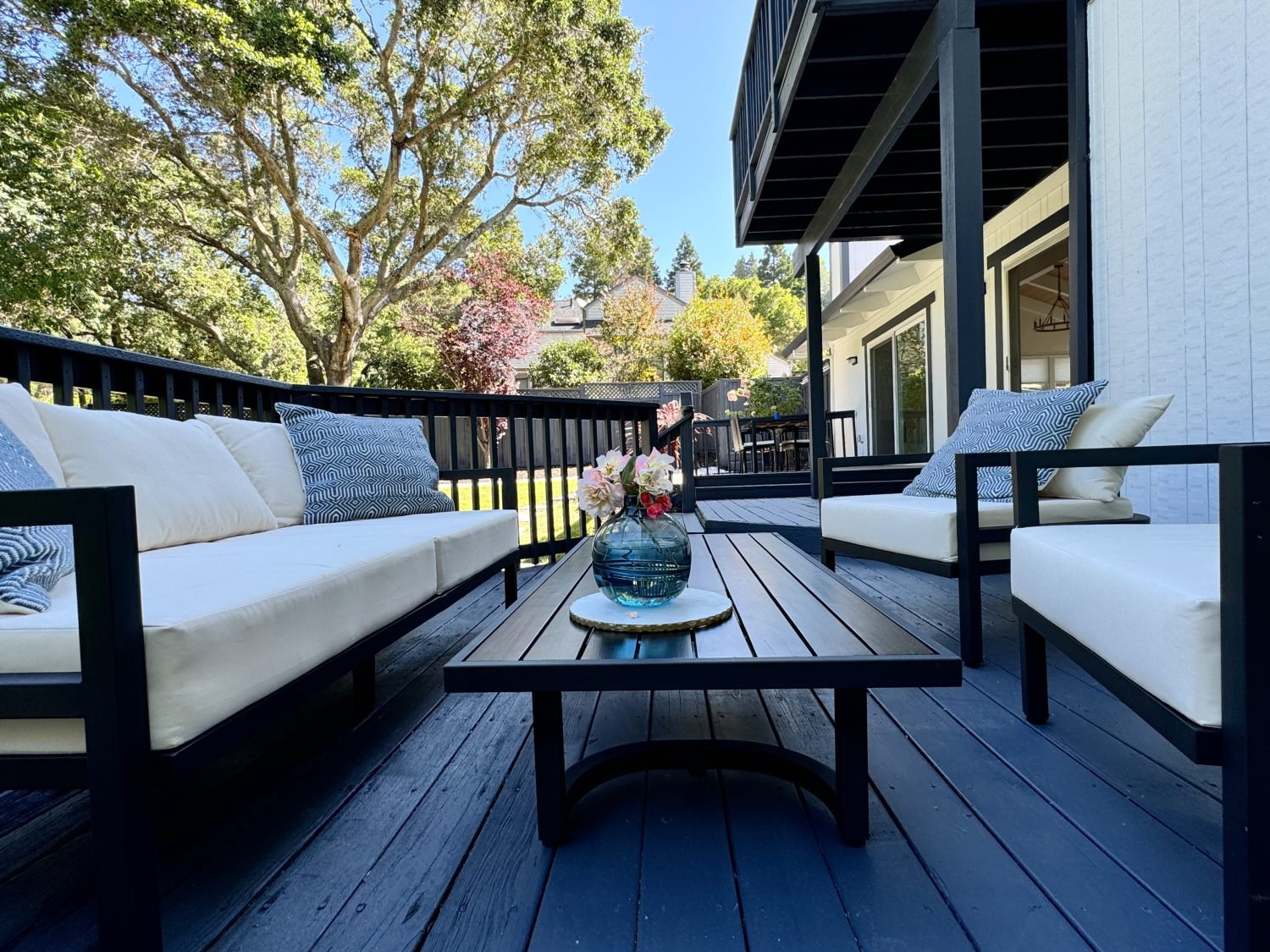 Outdoor patio with dark wooden decking, featuring white cushioned seating and a central coffee table with a vase of flowers. In the background, there are trees and a modern house exterior.