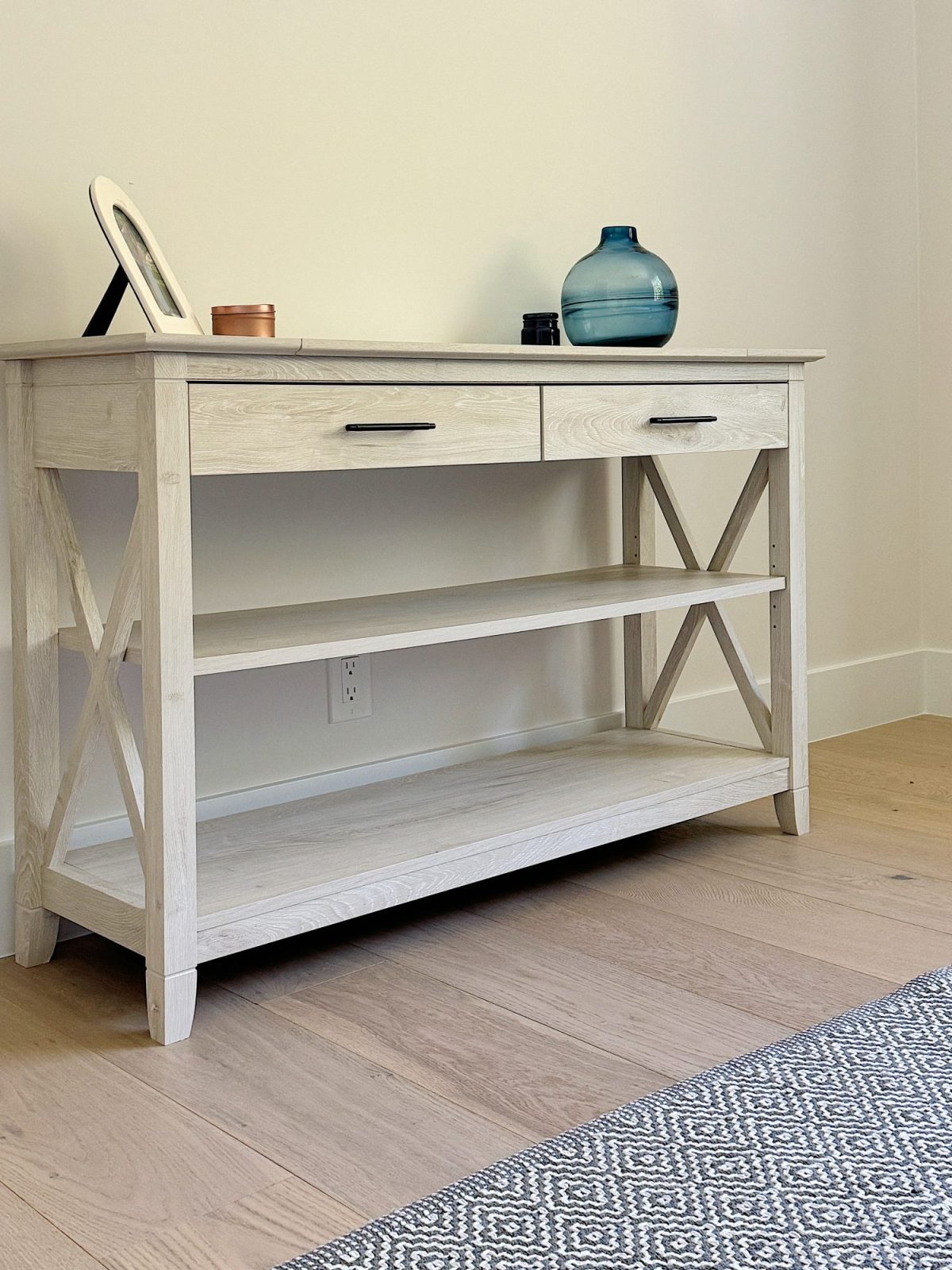 A white wooden console table with two drawers and two lower shelves, featuring a framed picture, a candle, and a blue glass vase on top, standing on a wooden floor near a patterned rug.
