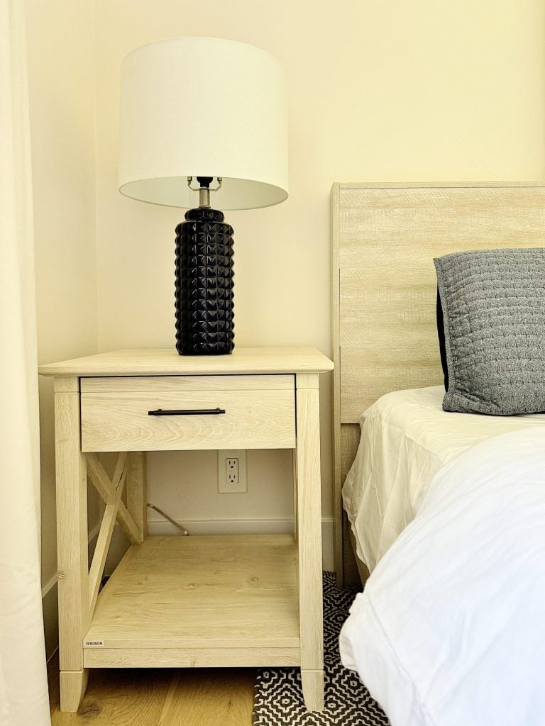 A bedside table with a drawer and shelf, featuring a black textured lamp with a white shade, a portion of a bed with a blue pillow, and a wall outlet beneath the table.