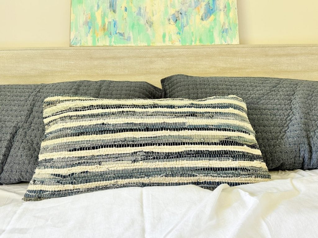 A bed with grey-blue knitted pillows, a striped cushion, and a colorful abstract painting above the headboard.
