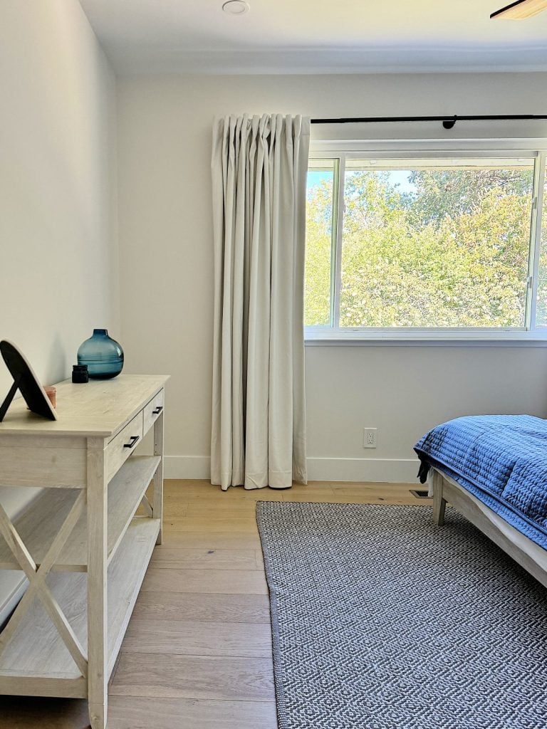 A minimalist bedroom with a light-colored dresser, a gray rug, a blue bedspread, a window with white curtains, and a view of trees outside.