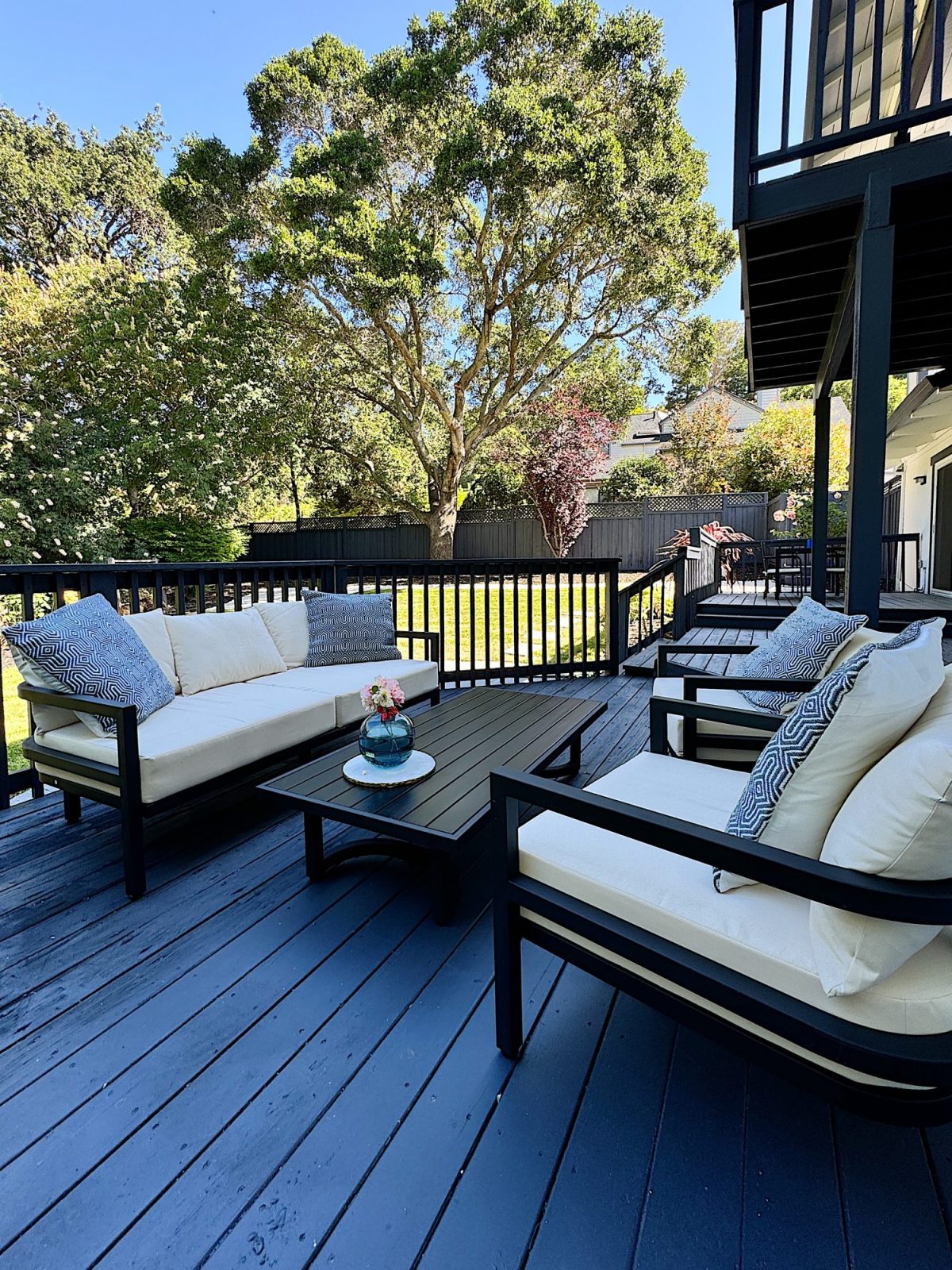 A backyard patio features white cushioned chairs, a small table with flowers, and surrounding greenery under a clear blue sky.