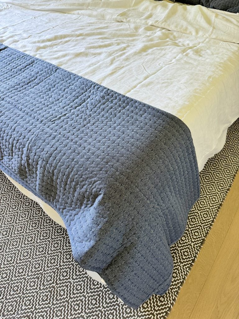 A bed with wrinkled white sheets, a blue quilt draped at the foot, and a patterned gray rug underneath.