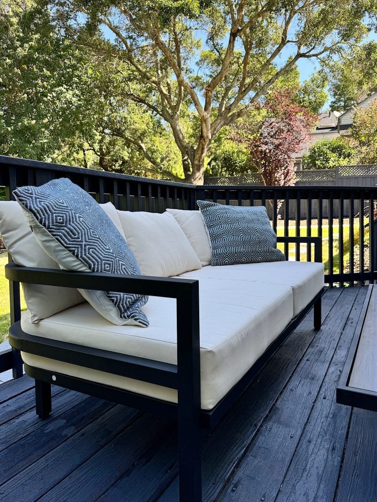 Outdoor patio featuring a sleek, black-framed couch with cream cushions and patterned pillows, set against a backdrop of a spacious yard with trees under a clear blue sky.