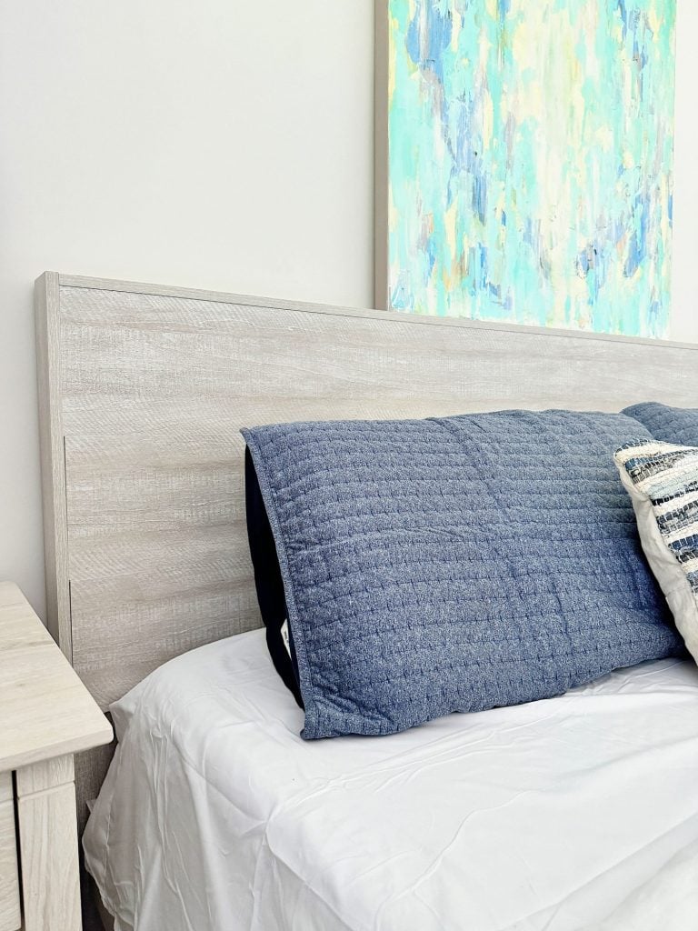 A bed with a light wood headboard, a white sheet, and blue pillows next to a bedside table with a colorful abstract painting hanging above.