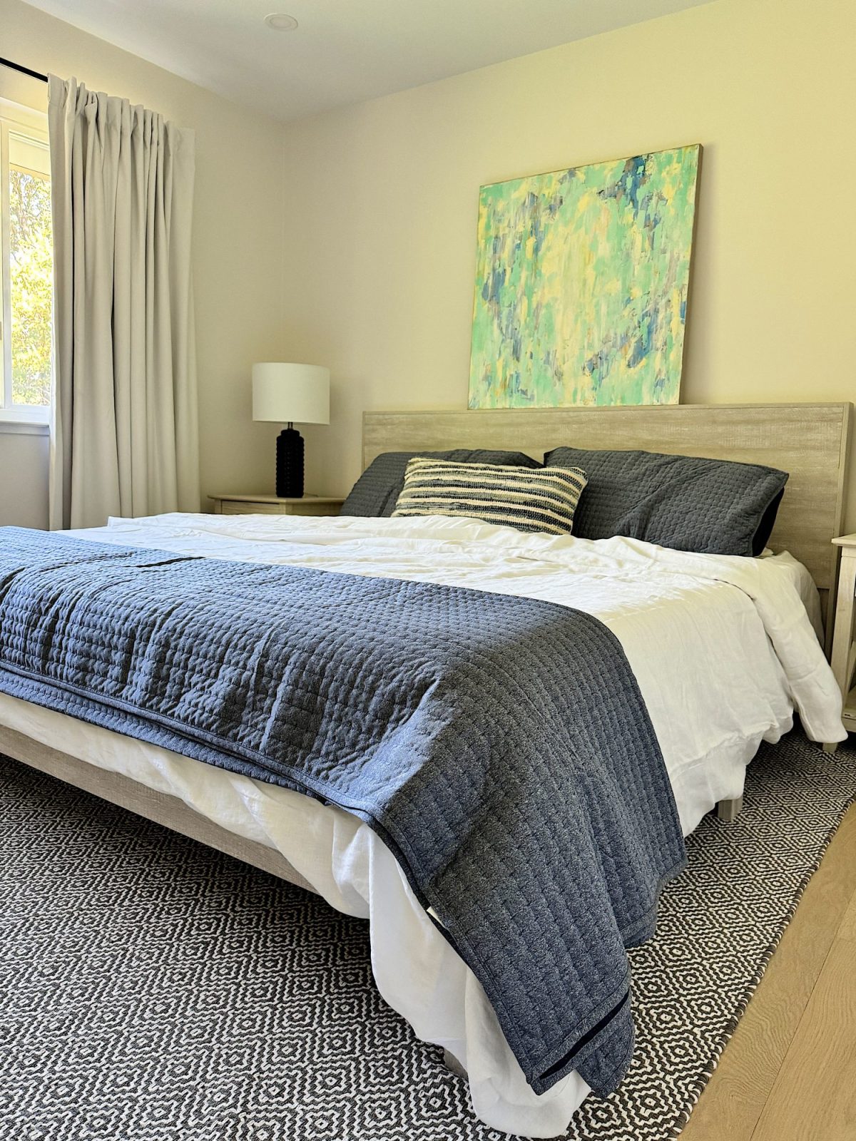 A bedroom with a neatly made bed featuring blue and white bedding, a bedside table with a lamp, a large abstract painting above the headboard, and light-colored curtains.