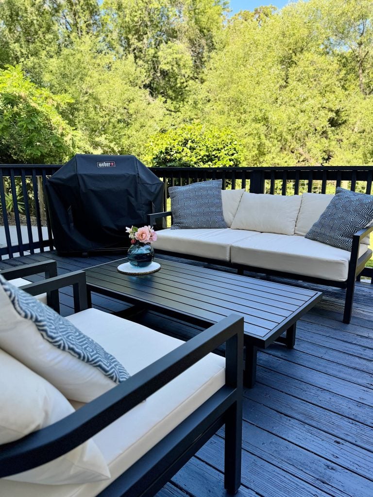 A patio with a black grill, white cushioned chairs, a black coffee table with a vase of flowers on a wooden deck, surrounded by trees.