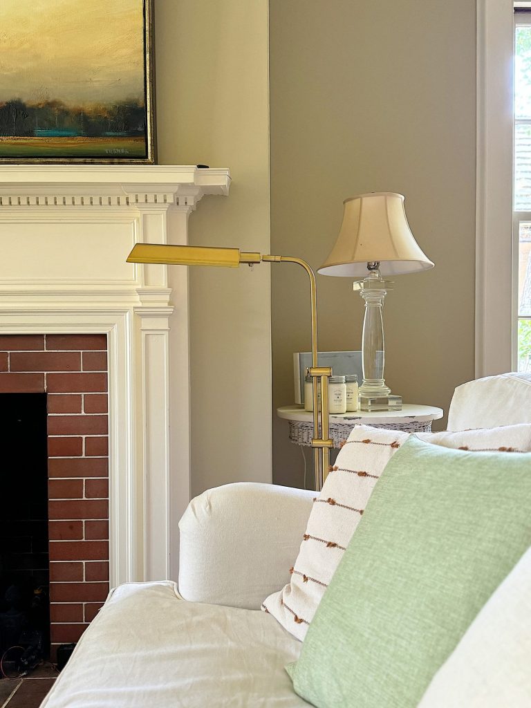 A cozy living room with a white fireplace and brick surround, a white couch with green and striped pillows, a brass floor lamp, and a side table with a lamp, books, and small decor items.