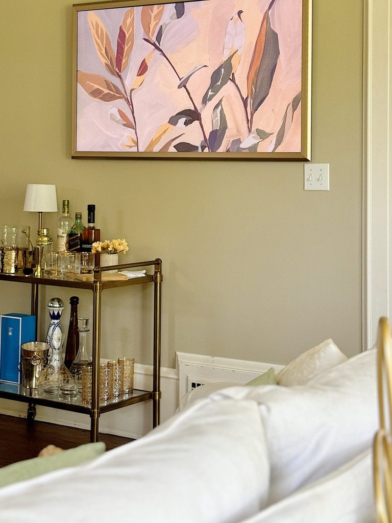 A wall-mounted painting of abstract leaves hangs above a brass bar cart stocked with bottles and glassware. A portion of a bed with light-colored bedding is visible in the foreground.