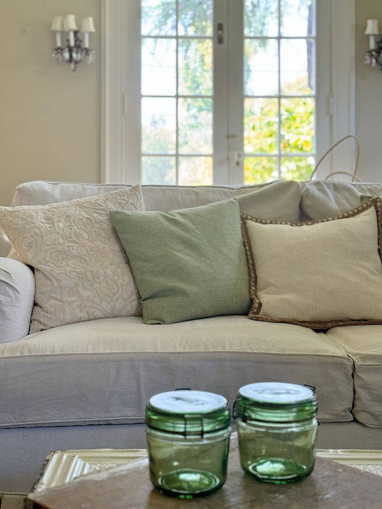 A beige sofa with various pillows, two green and three beige, sits in front of a window. A wooden table in the foreground holds two green glass jars.