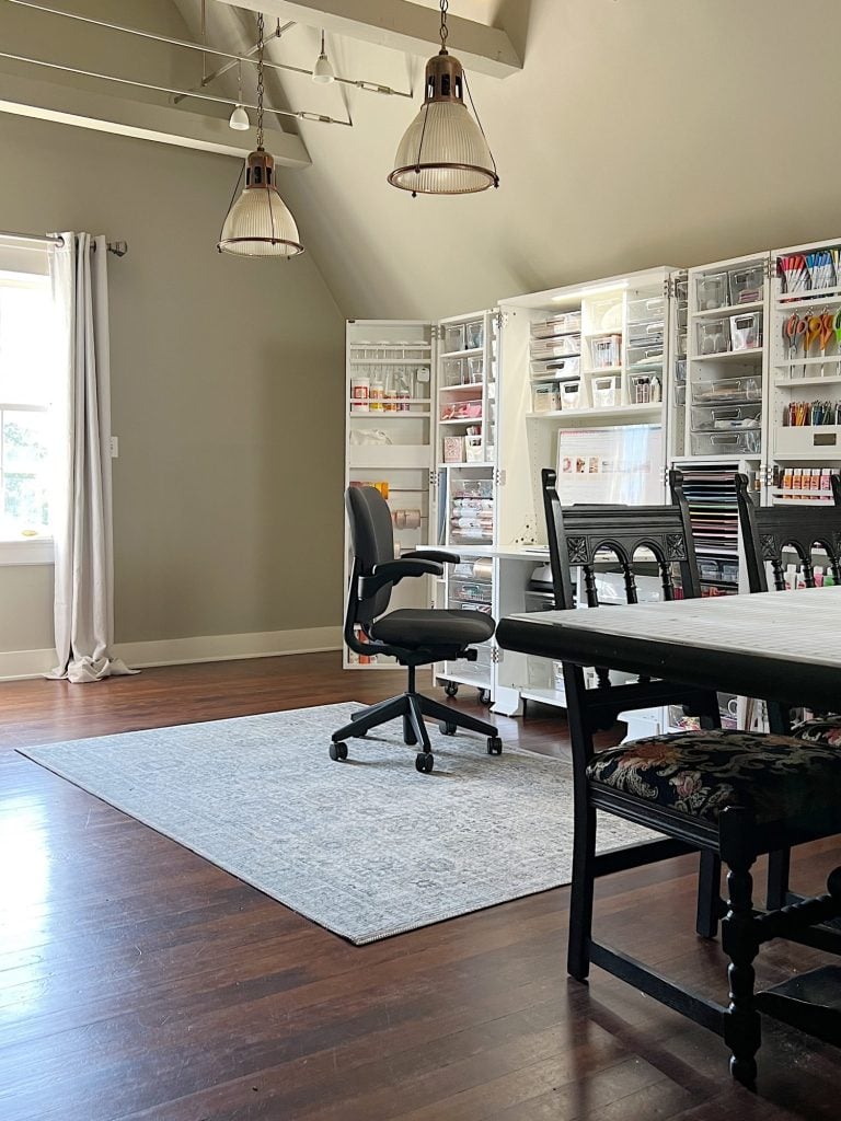 A well-organized craft room with wall-mounted storage units filled with supplies, a large work table, chairs, and a rolling office chair on a grey rug. Two pendant lights hang from the ceiling.