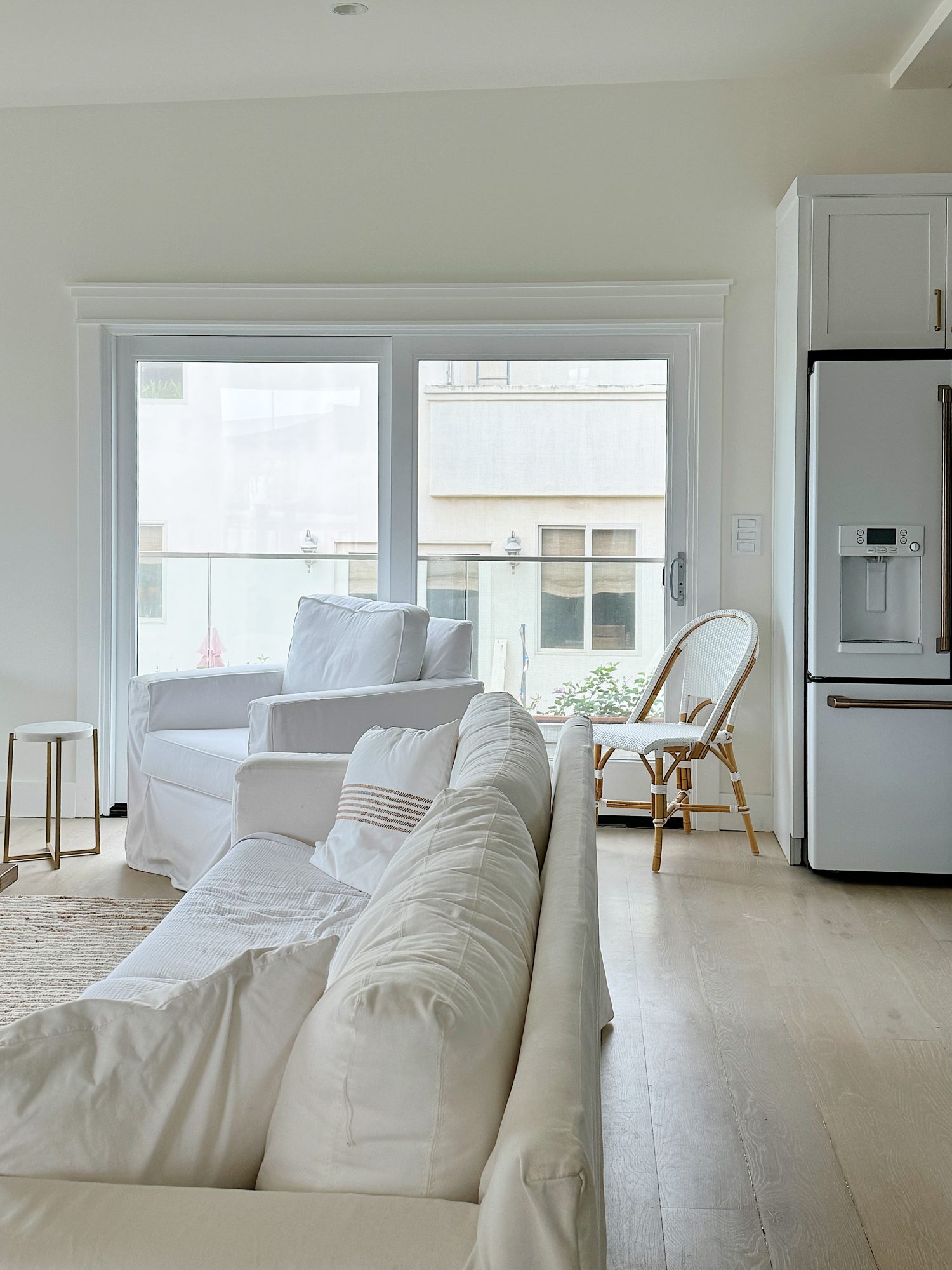 A bright living room with white furniture, including a sofa and an armchair, positioned near large windows. A rattan chair is placed next to a stainless steel refrigerator in the background.