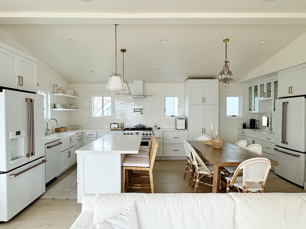 Modern kitchen with white cabinets, stainless steel appliances, an island with seating, and a wooden dining table with chairs. The space has pendant and chandelier lighting and an overall monochromatic white palette.