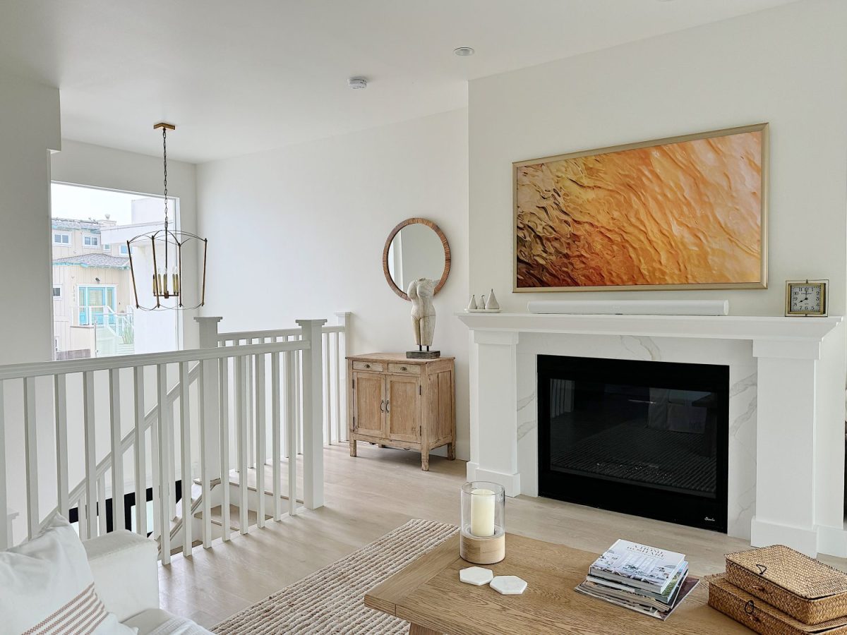 A modern living room with a white fireplace, a textured art piece, a wooden sideboard with a round mirror, and a staircase with white railings. A coffee table with decorative items is in the foreground.