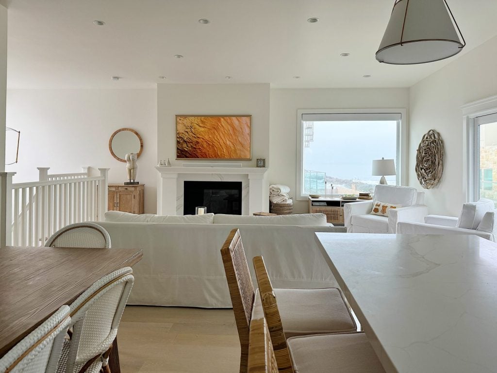 A cozy, modern living area with white furniture, a fireplace with a golden painting above, wicker chairs, and a large window offering a view of the sea. A wooden dining table and kitchen counter are visible.