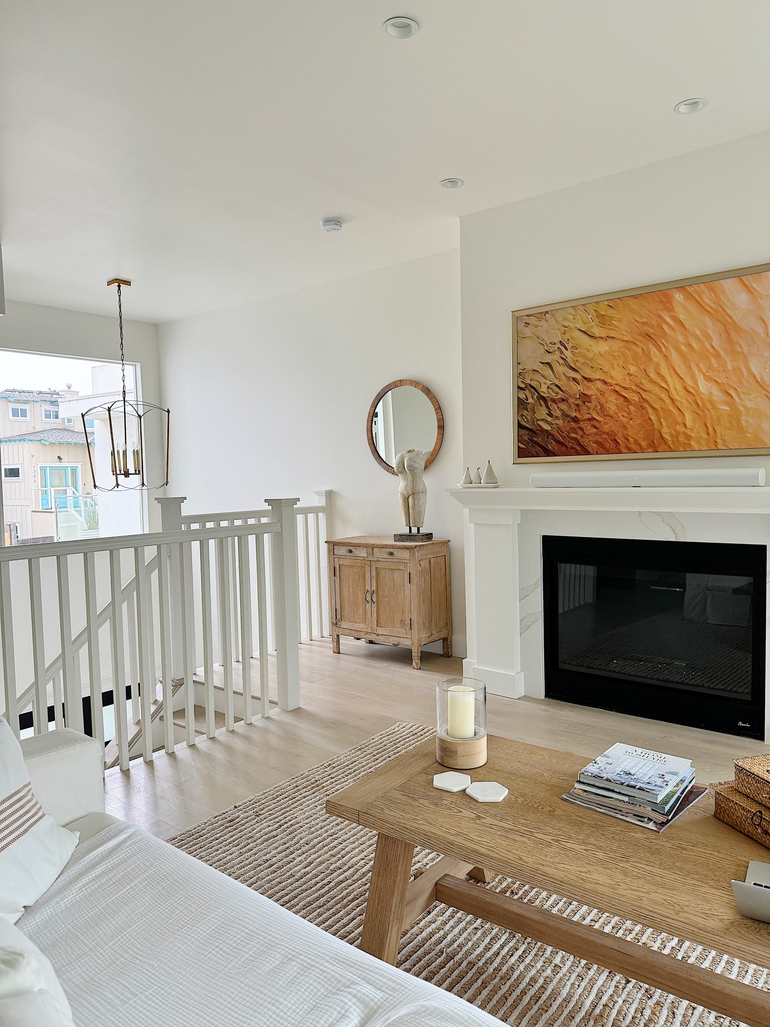 A modern living room features a white sofa, wooden coffee table, striped rug, fireplace with wall art above, wooden cabinet with a mirror, and a window with a view of buildings.