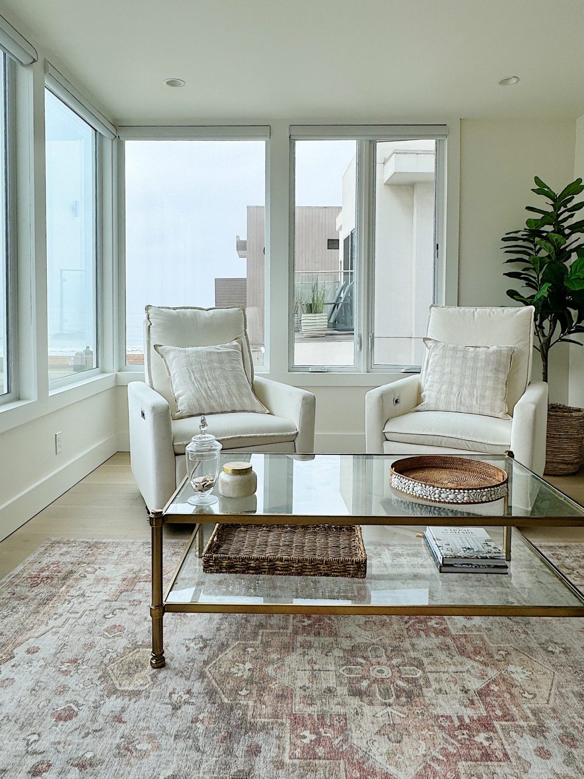 A modern living room with two white armchairs, a glass coffee table, a woven rug, and large windows allowing natural light to fill the space.