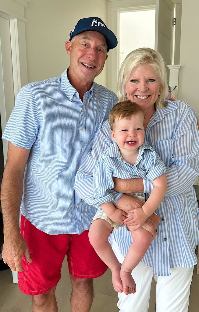 Two adults, one wearing a cap and red shorts, the other holding a smiling toddler, all dressed in light blue and white striped shirts, stand indoors near a door on their family vacation.