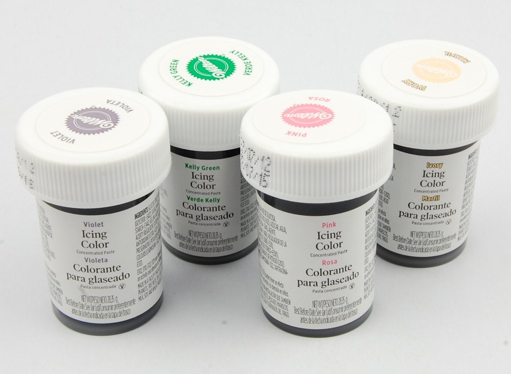 Four small jars of Wilton icing color in violet, kelly green, pink, and ivory—perfect for decorating treats like pink lemonade pie—each with white lids and labeled in English and Spanish, are arranged on a white surface.