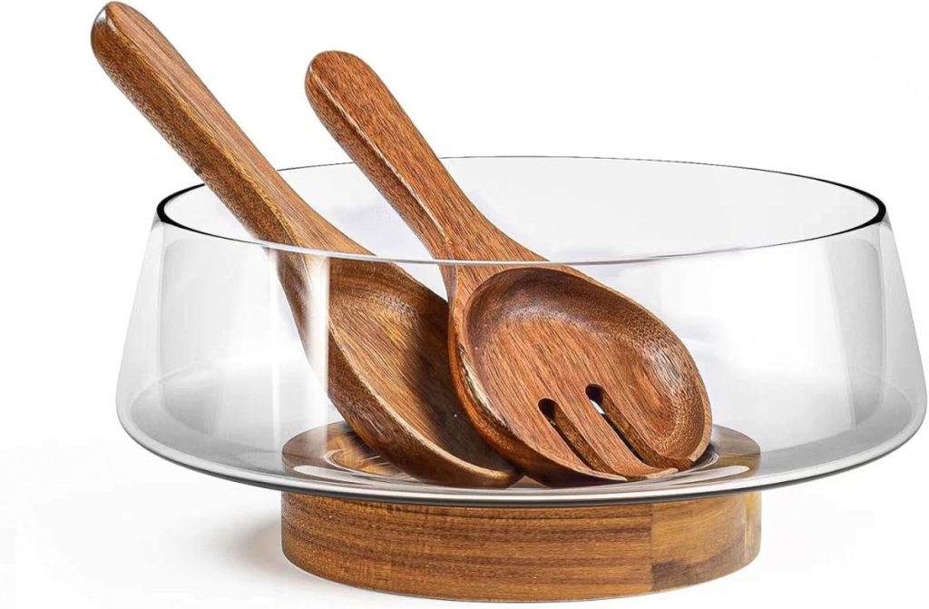 A clear glass salad bowl with a wooden base, containing a wooden serving spoon and fork—perfect for serving your favorite Summer Chicken Recipes in style.
