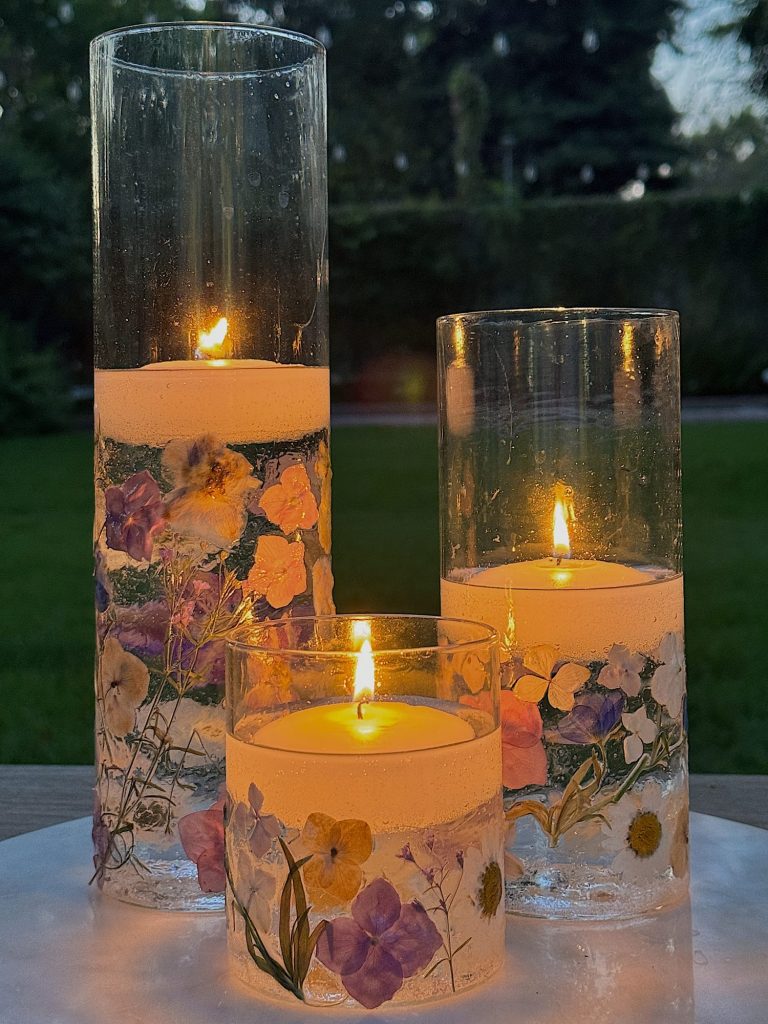 Three burning candles of varying heights, each encased in glass cylinders adorned with pressed flowers, placed on a white surface with a blurred green outdoor background.