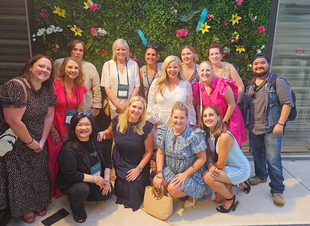 A group of fifteen people, including fourteen women and one man, pose for a photo in front of a wall decorated with green foliage and colorful flowers. They are smiling and dressed in various attires.