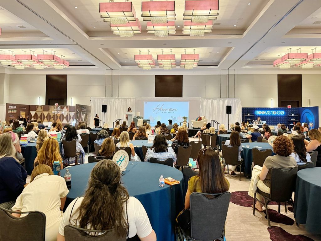 A conference room filled with seated attendees facing a stage where a presenter is speaking with a large screen behind them displaying text.