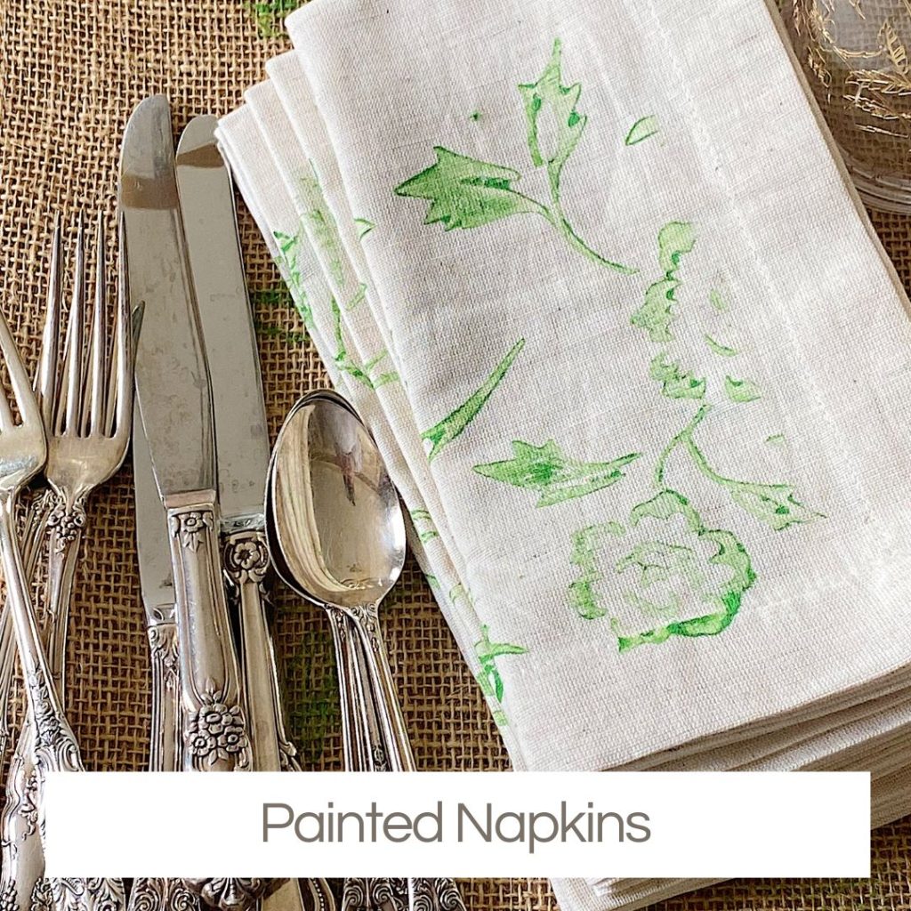 Silverware is arranged next to a folded stack of white napkins with green floral prints on a burlap surface. Text reads "Painted Napkins"—a charming addition to any Creative Crafts collection.