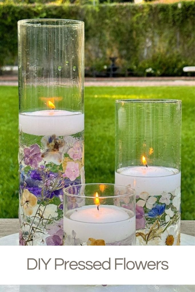 Three lit candles in tall cylindrical glass holders, each decorated with pressed flowers, placed on an outdoor table with grass and foliage in the background. Text at the bottom reads "DIY Pressed Flowers.