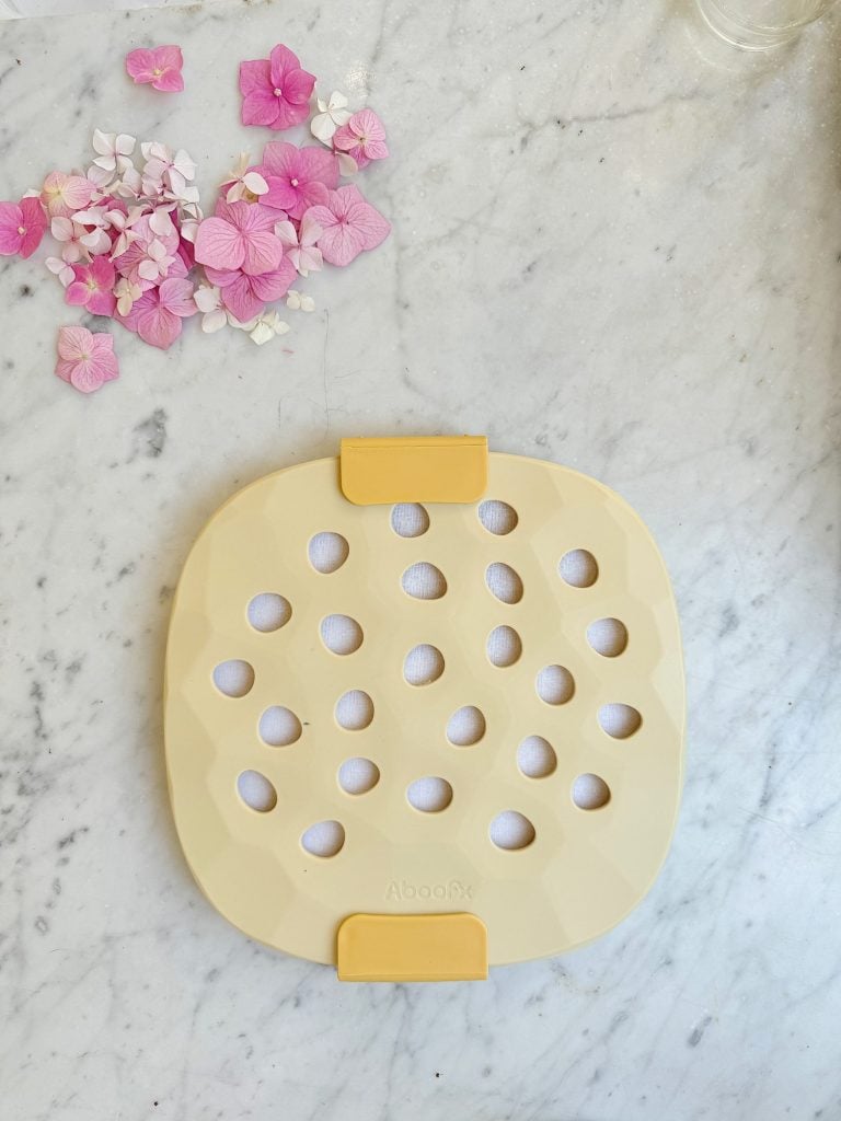 A beige silicone trivet with circular cutouts and yellow corners is placed on a white marble surface. Pink and white flower petals are scattered nearby.