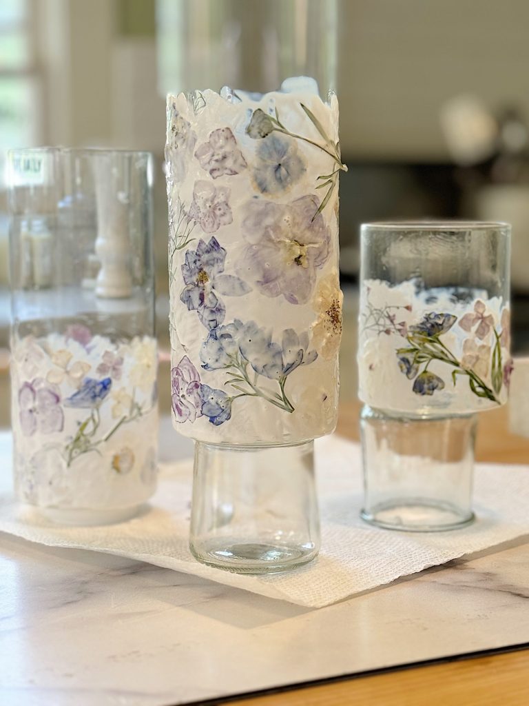 Three glass bottles decorated with pressed flowers and white plaster, arranged on a table covered with a white paper towel.