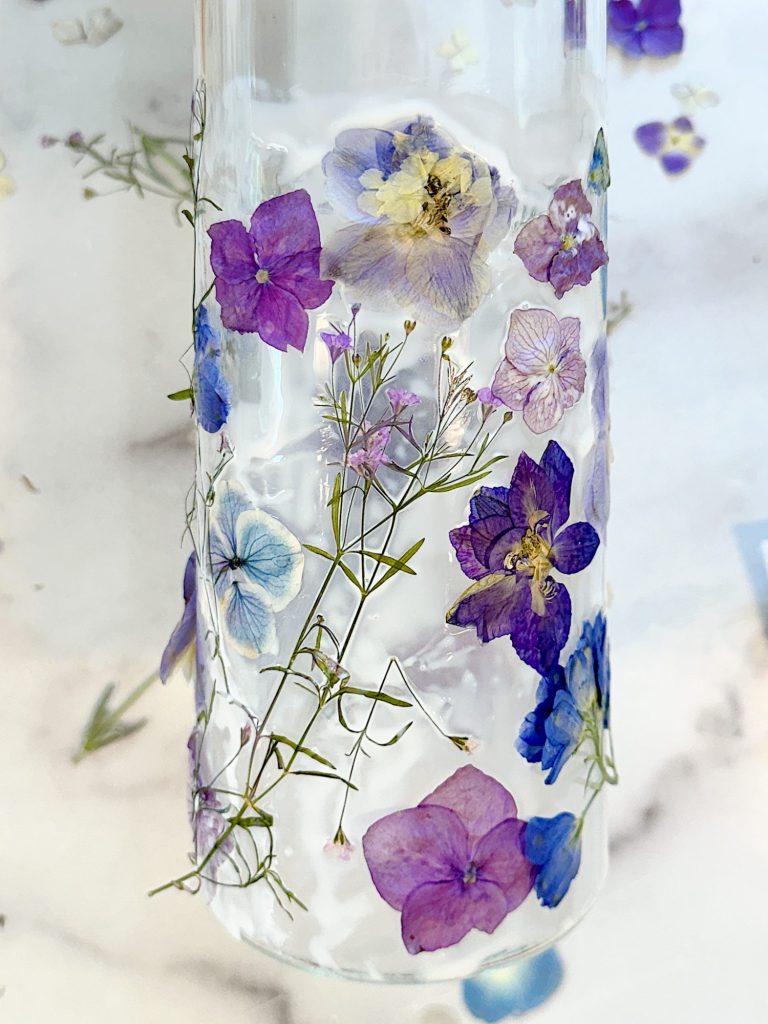 A clear bottle with pressed blue and purple flowers and green stems embedded inside. The bottle is placed on a surface with similar flower decorations.