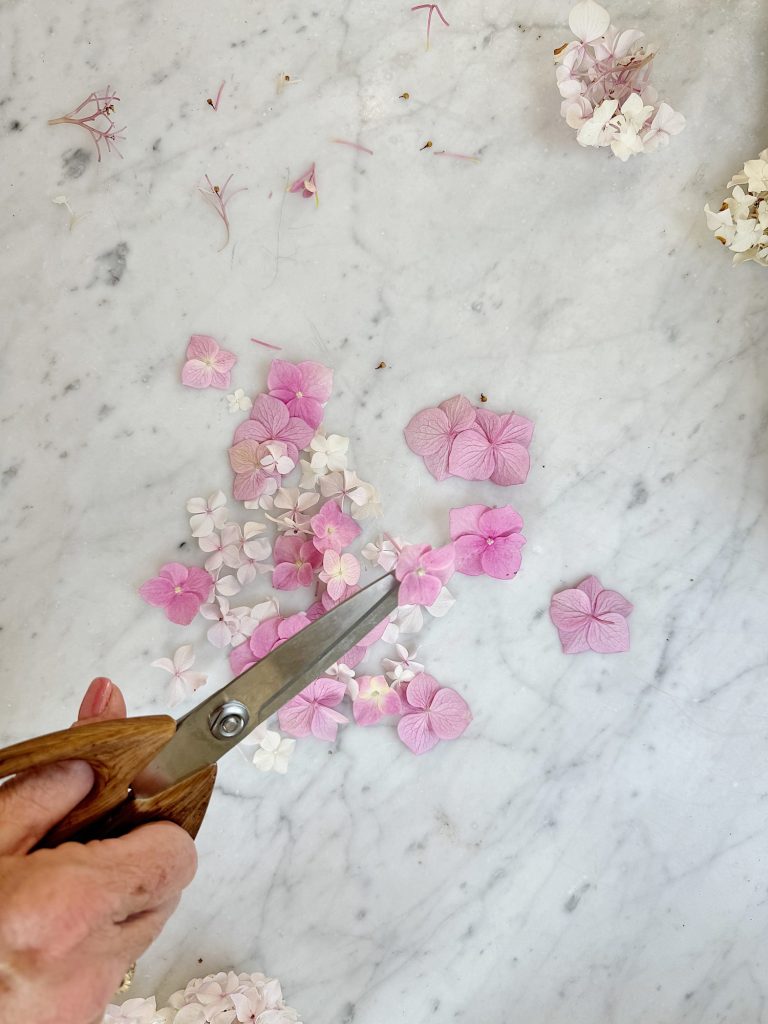 A hand holding scissors snips pink petals on a marble surface.