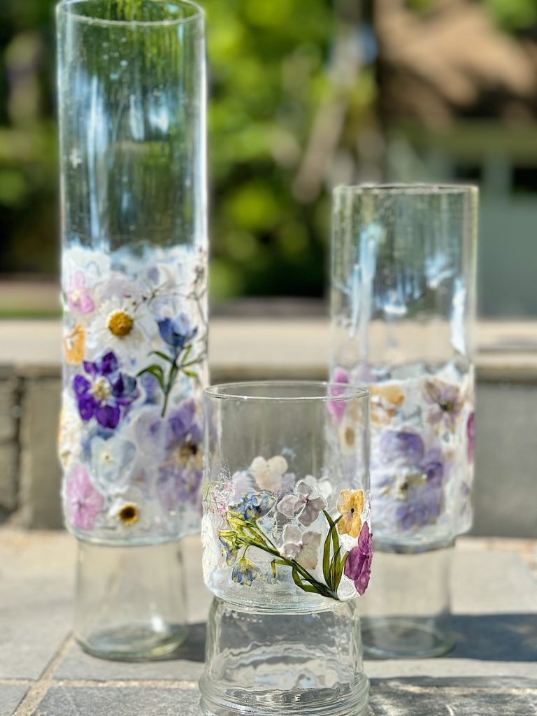 Three cylindrical glass vases of varying heights adorned with pressed flowers and foliage stand on a stone surface in an outdoor setting.