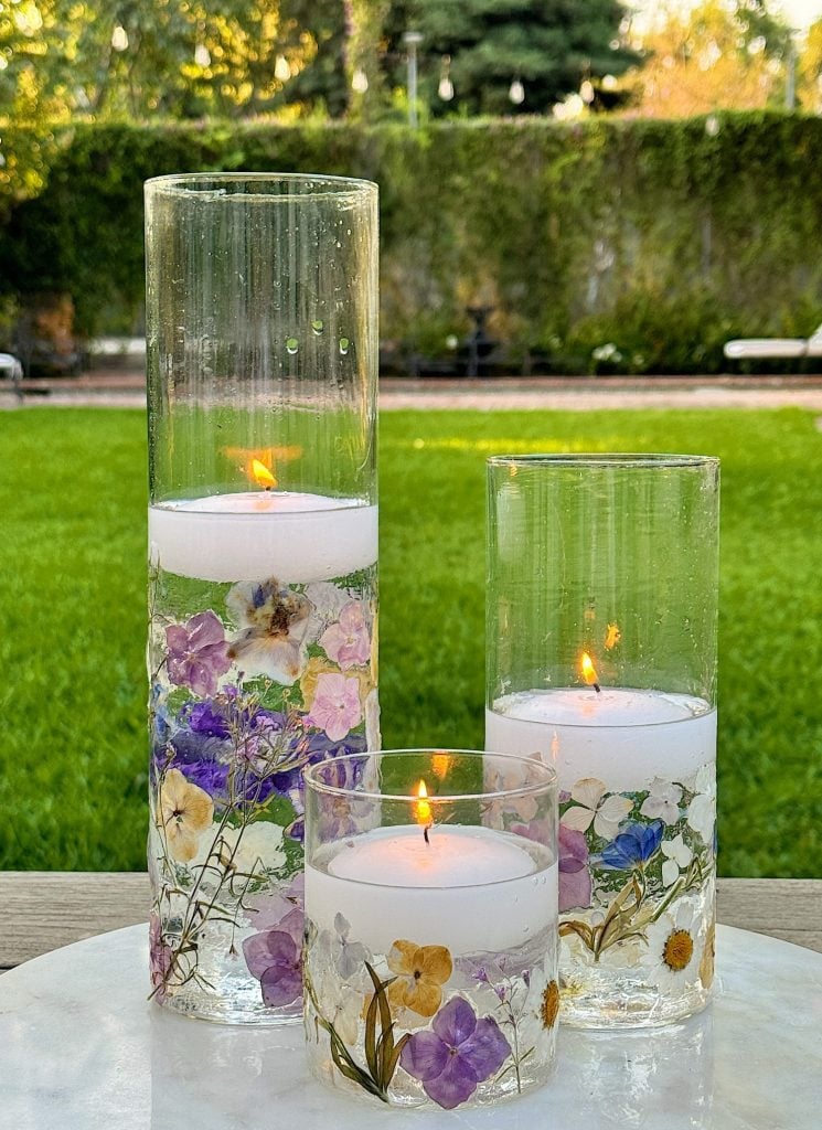 Three glass candle holders of varying heights with floating candles and dried flowers inside, set on a white surface outdoors with a green lawn and trees in the background.