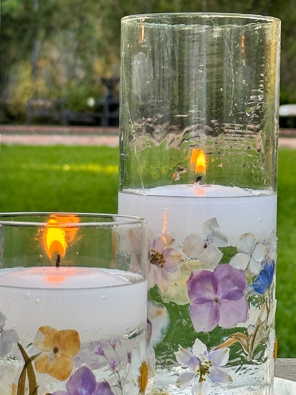 Two glass candle holders with lit white candles, adorned with pressed flowers, stand on an outdoor surface with grass in the background.