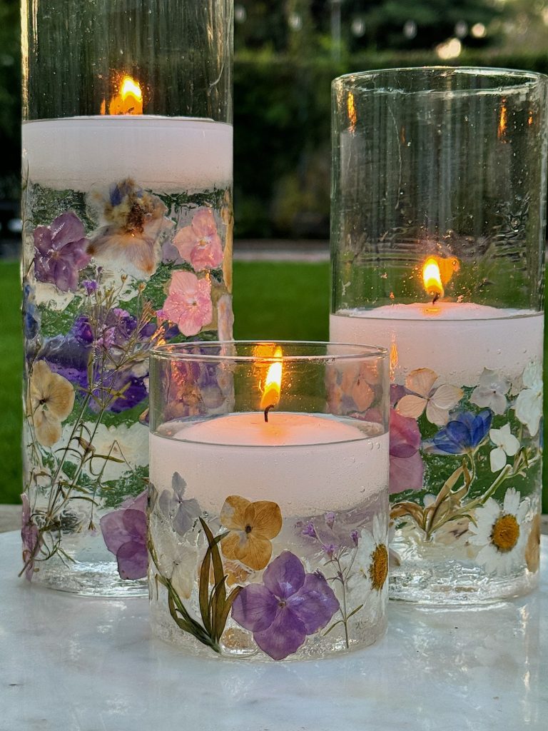 Three candles are lit inside clear cylindrical glass holders decorated with dried flowers. The holders are arranged on a white surface with a blurred green background.