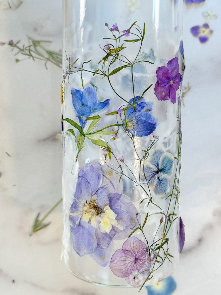 A glass vase filled with clear resin and embedded with various colorful, dried flowers and delicate green stems, displayed on a white surface with scattered flower petals.